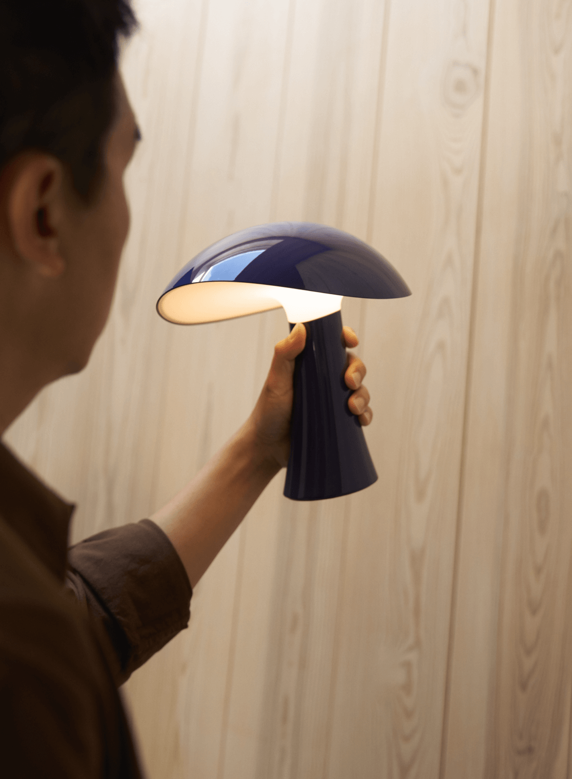 Person holding a Rumee portable lamp in a wooden interior, with soft lighting from the lamp, © 2025 Louis Poulsen